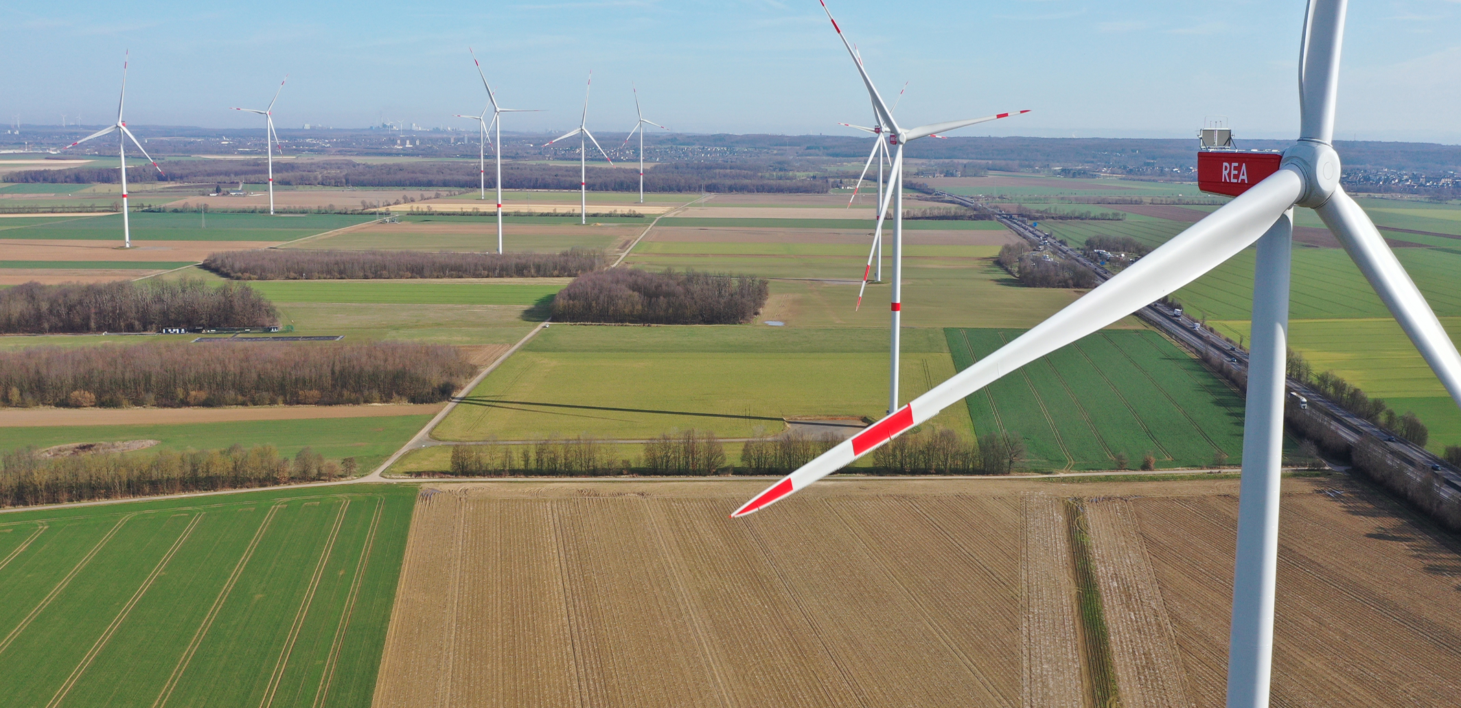 Luftaufnahme mehrerer Windkraftanlagen auf landwirtschaftlichen Flächen mit Feldern in verschiedenen Grün- und Brauntönen unter einem klaren blauen Himmel. Eine Turbine ist deutlich im Vordergrund zu sehen.