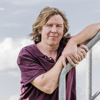 Ein Mann mit mittellangem hellbraunem Haar und kastanienbraunem Polohemd lehnt im Freien an einem Metallgeländer, im Hintergrund ist der Himmel bewölkt.