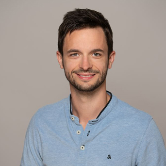 Ein Mann mit kurzen dunklen Haaren und einem gestutzten Bart lächelt. Er trägt ein hellblaues langärmeliges Hemd mit Knöpfen am Kragen. Der Hintergrund ist einfarbig und hell.