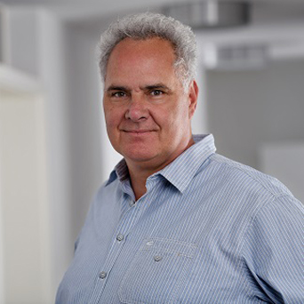 Ein Mann mittleren Alters mit kurzen, gelockten grauen Haaren und einem hellblauen Hemd zum Aufknöpfen steht in einem Haus und schaut mit neutralem Blick in die Kamera. Der Hintergrund ist sanft verschwommen.