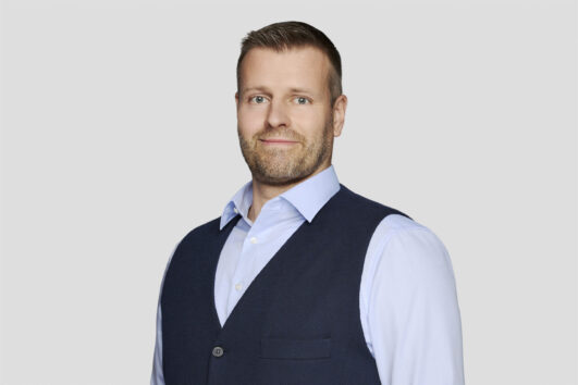 Ein Mann mit kurzen blonden Haaren und einem Bart trägt ein hellblaues Hemd und eine dunkelblaue Weste und steht vor einem schlichten hellgrauen Hintergrund.